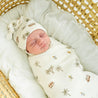 A newborn baby, swaddled in an Organic Swaddle Blanket & Hat with palm tree print, is asleep in a woven basket, wearing a matching beanie from Makemake Organics.