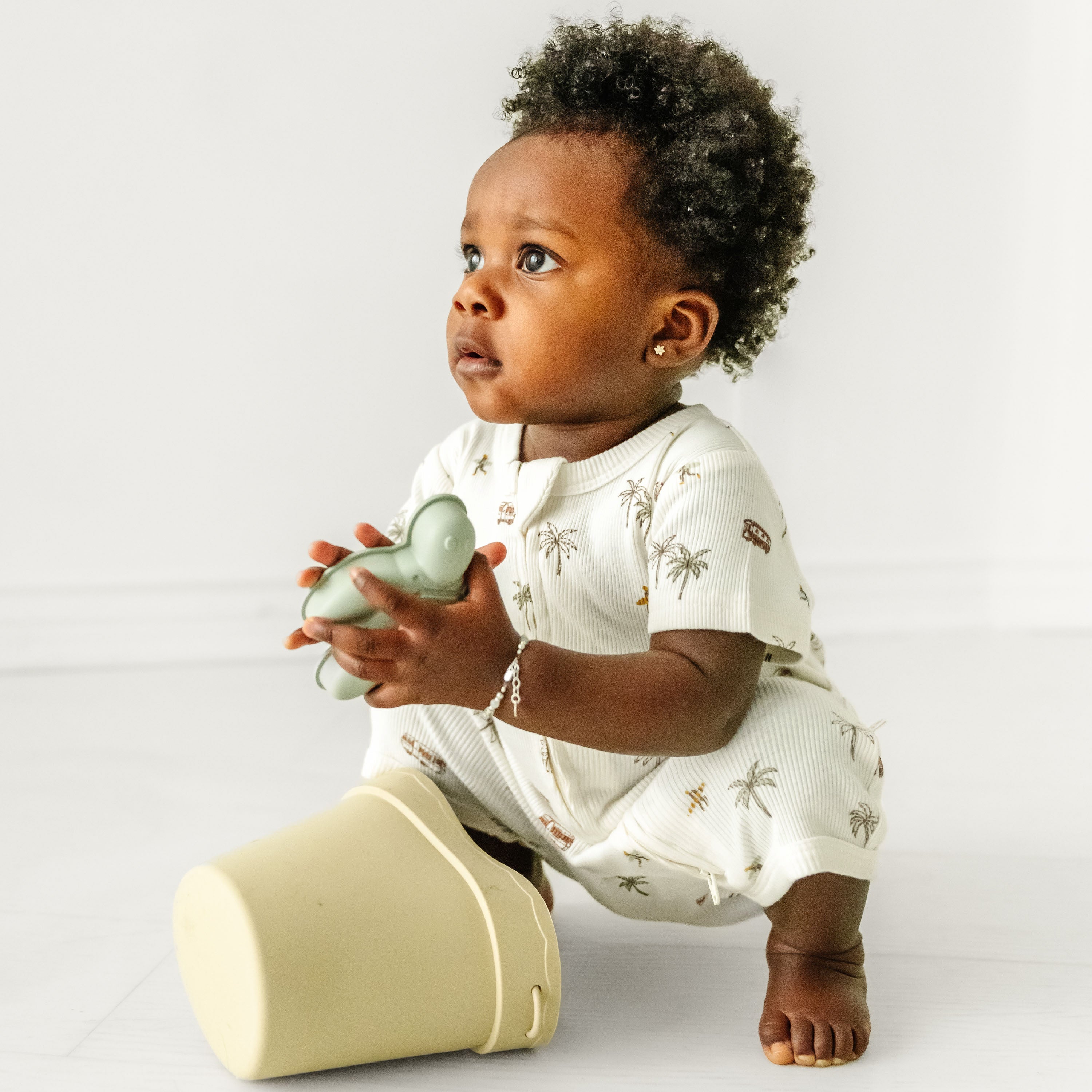 A baby with curly hair, wearing a white Makemake Organics Organic Short Zip Romper with animal prints, holds a toy and sits on a white floor, looking thoughtfully to the side.