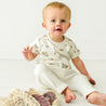 A surprised-looking baby sits on the floor, wearing a cream-colored onesie with a nature-inspired print, next to a wicker basket holding the Organic Tee & Pants Set from Makemake Organics.