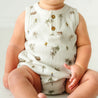 A toddler in an Organic Bubble Onesie by Makemake Organics with a tropical pattern sits on a bright surface, showing only from the stomach down. The focus is on the chubby hands and legs.
