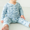 A toddler wearing a blue and white feather-patterned Organic Kimono Top & Pants Set from Makemake Organics sits on a white floor, with a partial focus on their tiny feet.