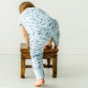 A toddler girl in a blue and white patterned Organic Short Sleeve Button Romper from Minnow by Makemake Organics is bending over, trying to climb onto a small, rustic wooden stool, against a plain white wall.