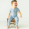 A joyful toddler boy in a Minnow Organic Short Sleeve Button Romper sits on a rustic wooden stool against a plain background, smiling broadly. (Brand Name: Makemake Organics)