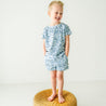A young child in a Organic Tee and Shorts Set from Minnow, smiling and standing barefoot on a woven stool against a plain white background.