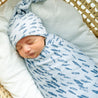 Newborn baby wrapped in a blue Organic Swaddle Blanket with fish patterns and wearing a matching hat, sleeping peacefully in a woven basket from Makemake Organics.