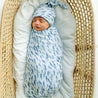 A newborn baby wrapped in a Minnow Organic Swaddle Blanket and Hat, sleeping peacefully in a wicker bassinet lined with a white cushion by Makemake Organics.