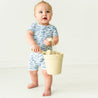A toddler boy in a blue and white outfit stands barefoot, holding a cream-colored bucket by its handle, looking slightly surprised in a bright room wearing the Organic Short Zip Romper by Makemake Organics.