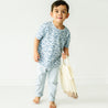 A young boy in a blue and white fish-patterned shirt and blue pants, standing barefoot and holding a white mesh bag, smiles in a bright, simple studio setting wearing the Organic Tee & Pants Set by Minnow from Makemake Organics.