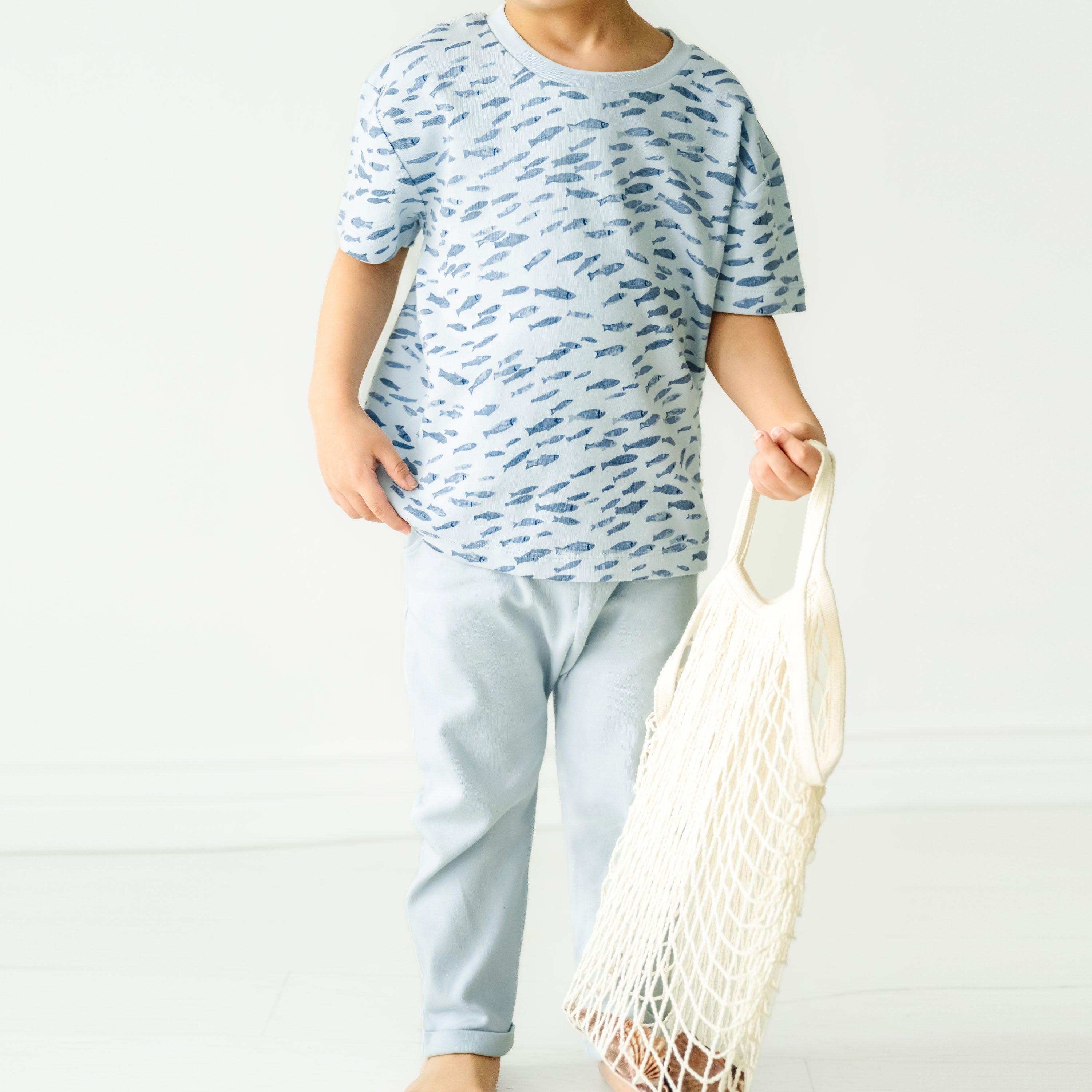 A child in casual blue attire, holding an empty white mesh bag, standing in front of a plain white background. only the torso and legs of the child are visible wearing an Organic Tee & Pants Set by Minnow from Makemake Organics.