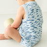 A baby in an Organic Bubble Onesie by Makemake Organics patterned with white fish sits on the floor, facing away from the camera and holding a small yellow object.