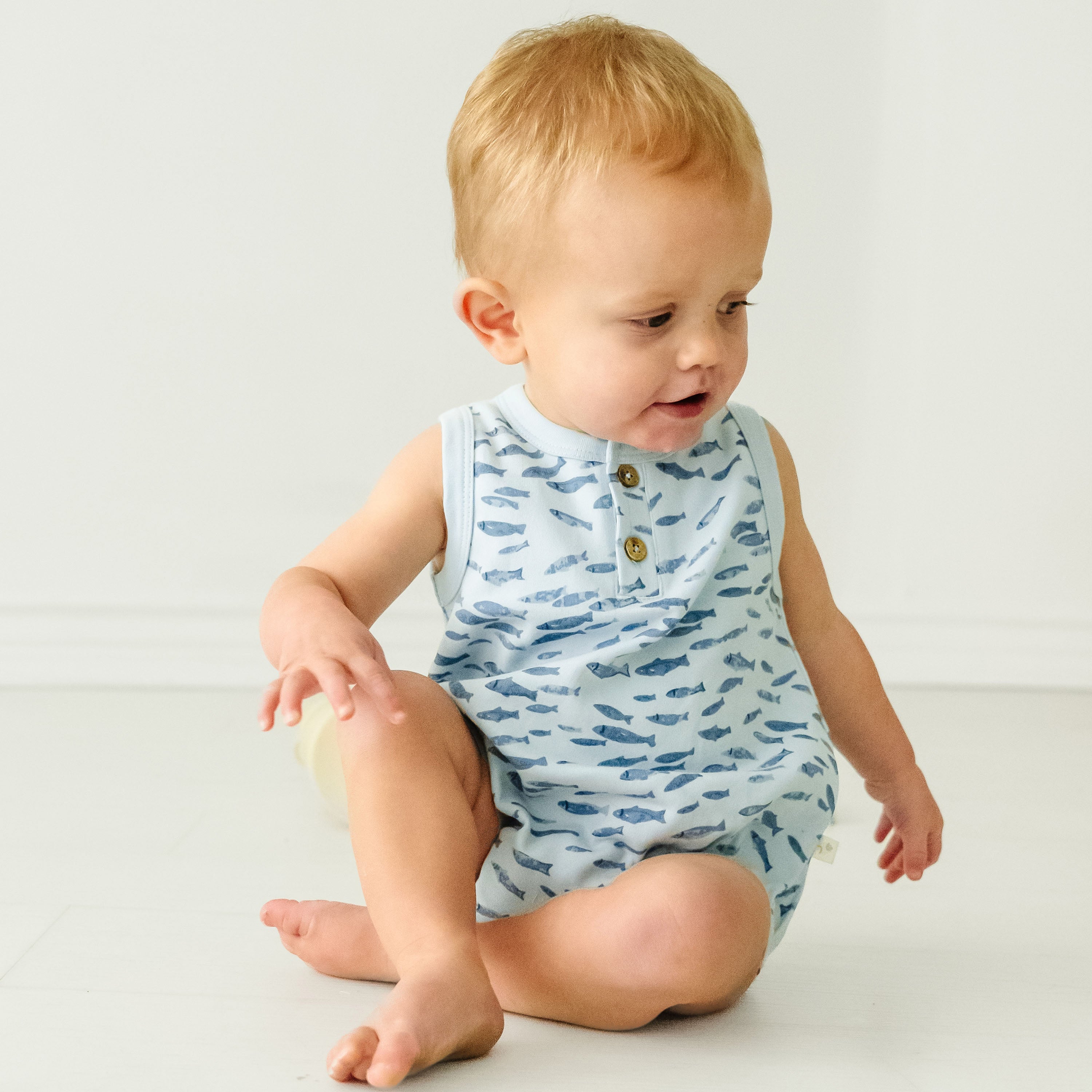 A baby with blond hair in a blue fish-patterned Minnow Organic Bubble Onesie from Makemake Organics sits on the floor, looking to the side with an expression of curiosity.