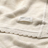Close-up view of a cream-colored, ribbed, Makemake Organics Organic Cotton Scalloped Baby Blanket with a "Makemake Organics" label attached at the seam.