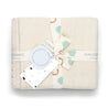 Replace with: A neatly packaged Makemake Organics Organic Cotton Scalloped Baby Blanket in Vanilla Natural with a pattern of teardrop shapes in shades of teal and beige, tied with a ribbon and a decorative tag.