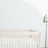 A minimalist nursery with a Makemake Organics light wood crib adorned with a Dino Park patterned bedding set, standing next to a floor lamp. The room has a gentle blue half-circle painted on a white wall.