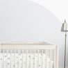 A minimalist nursery room featuring a white Makemake Organics crib with patterned bedding and a metallic floor lamp, set against a partially painted grey and white wall.
