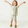 A joyful toddler with arms raised wide, smiling in a studio. She wears an Organic Linen Flutter Top and Shorts - Palms by Makemake Organics, and brown shoes against a plain white background.