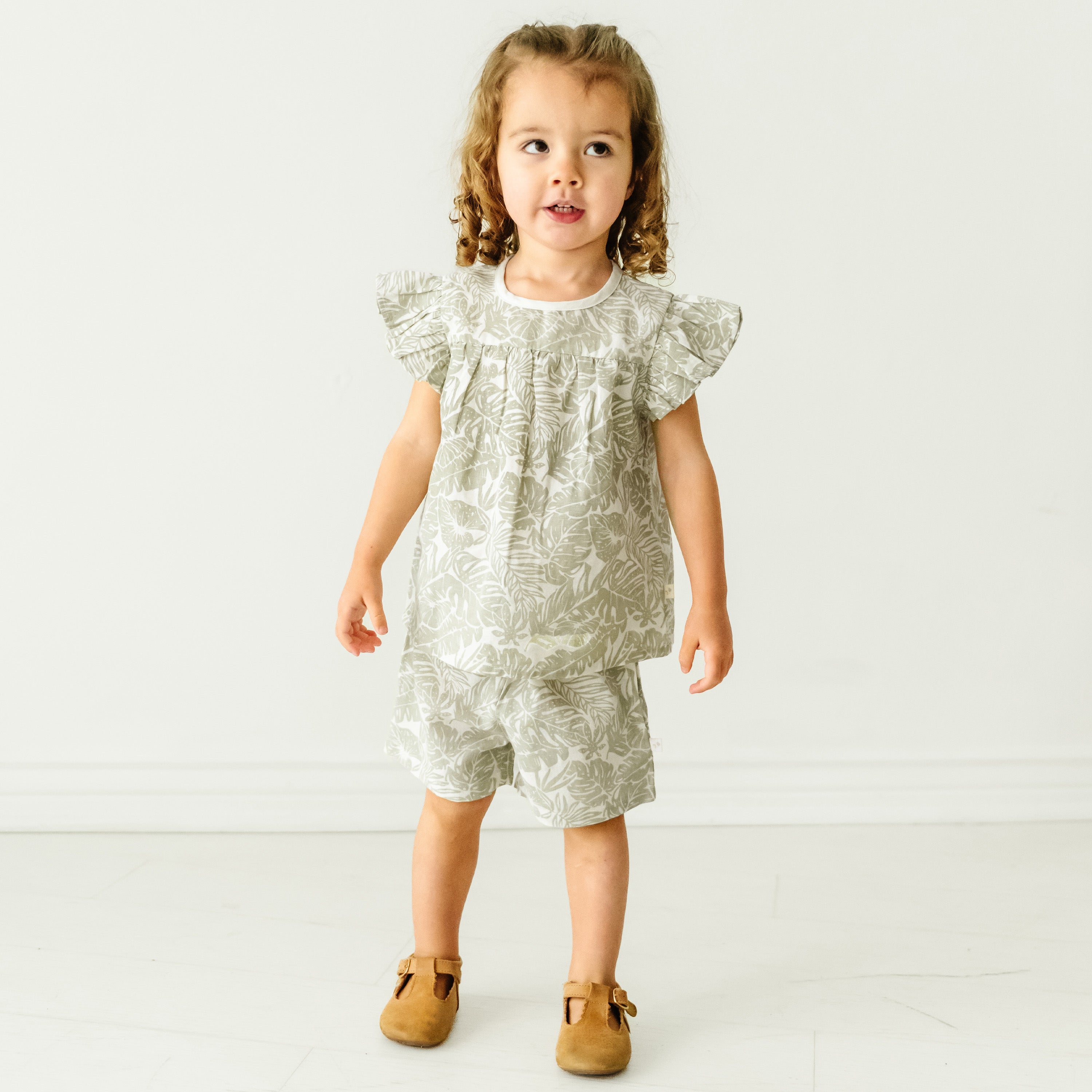 A toddler boy with curly hair stands smiling in a Makemake Organics Organic Linen Flutter Top and Shorts - Palms and tan shoes against a plain white background.