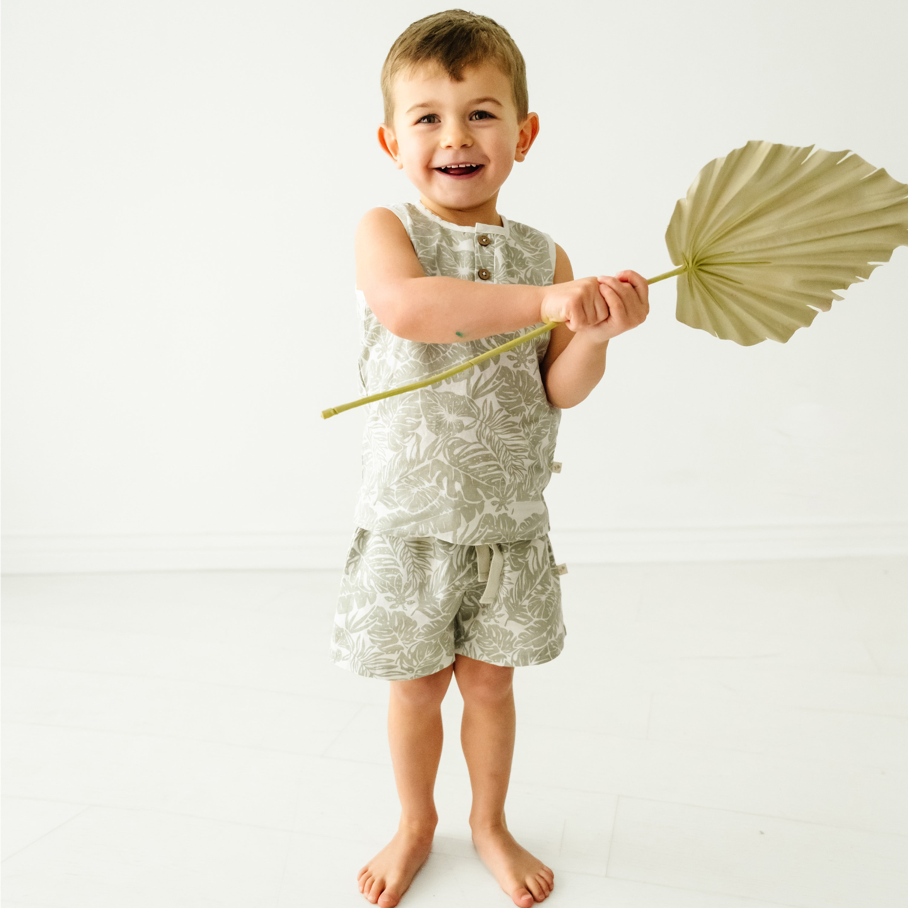 A cheerful young girl in a Makemake Organics Organic Linen Tank and Shorts Set - Palms smiles widely, holding a large palm leaf against a plain white background.
