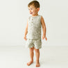 A toddler stands barefoot, smiling in a studio setting, dressed in a Makemake Organics Organic Linen Tank and Shorts Set featuring a Palms leaf pattern. He appears to be mid-conversation or reacting to something amusing.