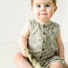 A toddler boy with wide blue eyes and light skin wearing a Organic Linen Sleeveless Bubble Romper in Palms print by Makemake Organics, sitting on a plain light background.