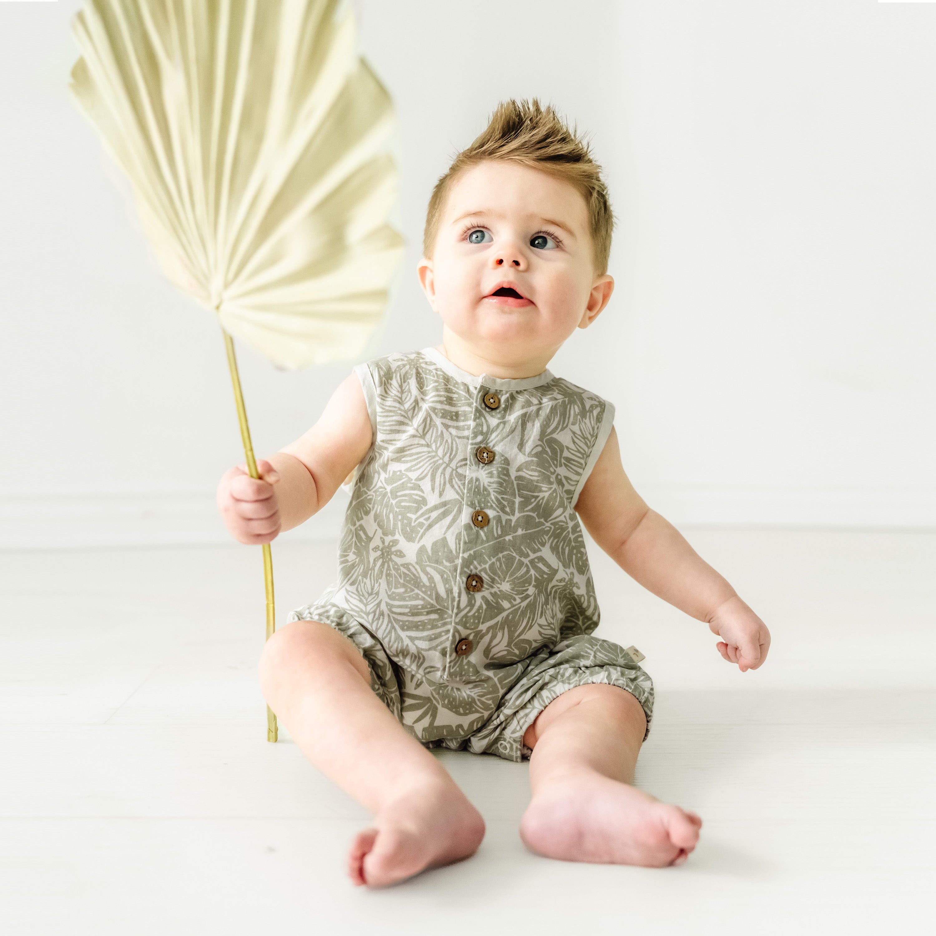 A toddler girl with a spiky hairstyle sits on a white floor, holding a golden leaf-shaped fan, wearing an Organic Linen Sleeveless Bubble Romper with leaf patterns from Makemake Organics.