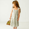 A young girl in a Organic Linen Tiered Strap Dress - Palms by Makemake Organics, holding a wicker basket, standing against a plain white background and looking away to the side.