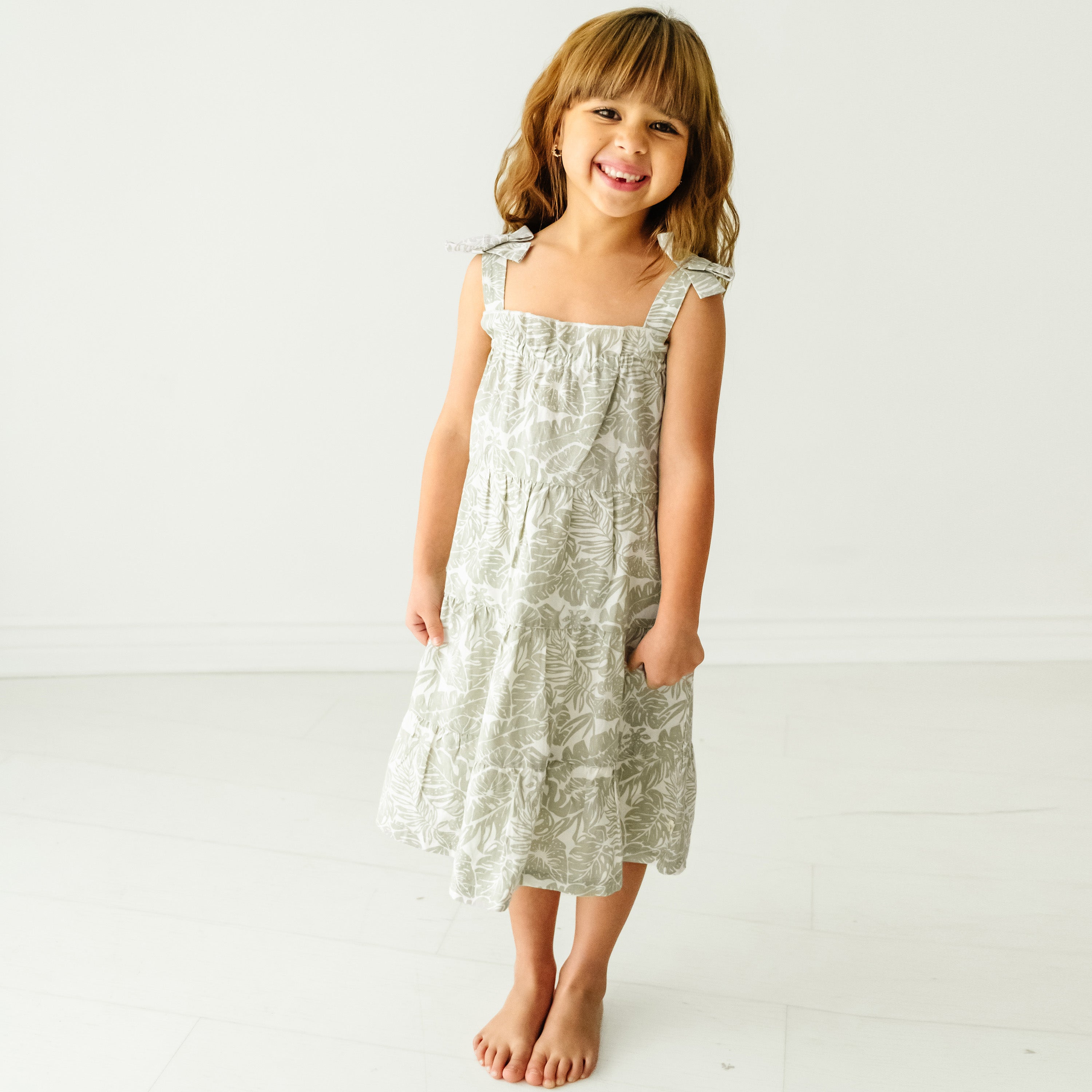 A young girl with shoulder-length brown hair smiles at the camera, wearing a white and green floral print dress. She is standing barefoot in a bright, neutral-toned room. The dress she is wearing is the Organic Linen Tiered Strap Dress in Palms by Makemake Organics.
