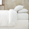A neatly made bed with white bedding and striped pillows against a plain gray wall. the bed includes a Organic Cotton Sheet Set - Pebble from Makemake Organics, which consists of a fitted sheet, duvet, and four pillows arranged in layers.
