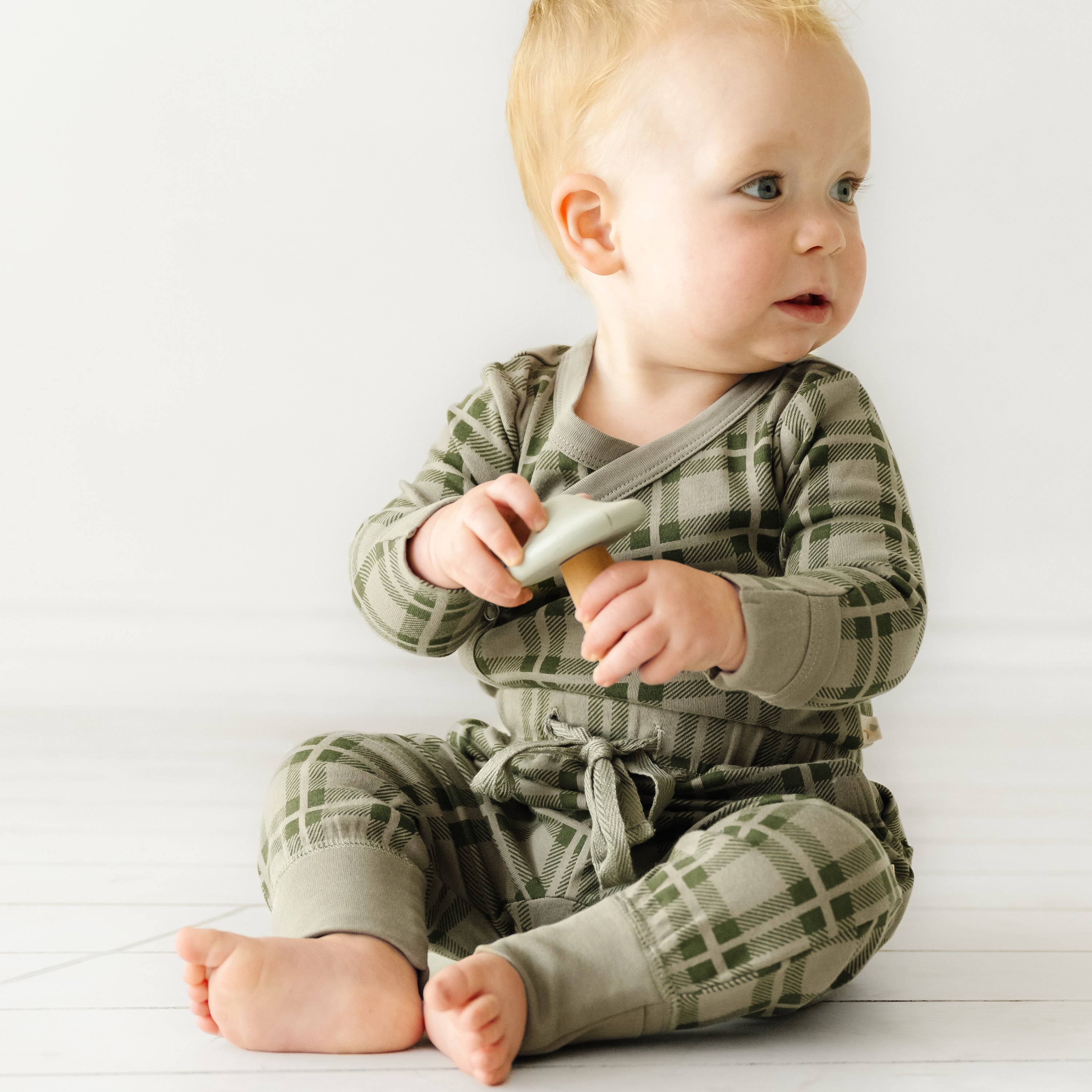 A baby with light hair sits on a white floor, wearing an Organic Baby green checkered outfit and holding a toy, looking to the side with a curious expression.