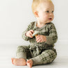 A baby with light hair sits on a white floor, wearing an Organic Baby green checkered outfit and holding a toy, looking to the side with a curious expression.