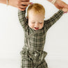 A toddler with light hair, dressed in an Organic Baby checkered jumpsuit, takes assisted steps while holding onto an adult's hands, smiling joyfully against a white background.
