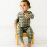 A toddler with curly hair sits on a small wooden chair, wearing an Organic Baby green and white plaid 2-Way Zip Romper, reaching out playfully. The background is plain white.
