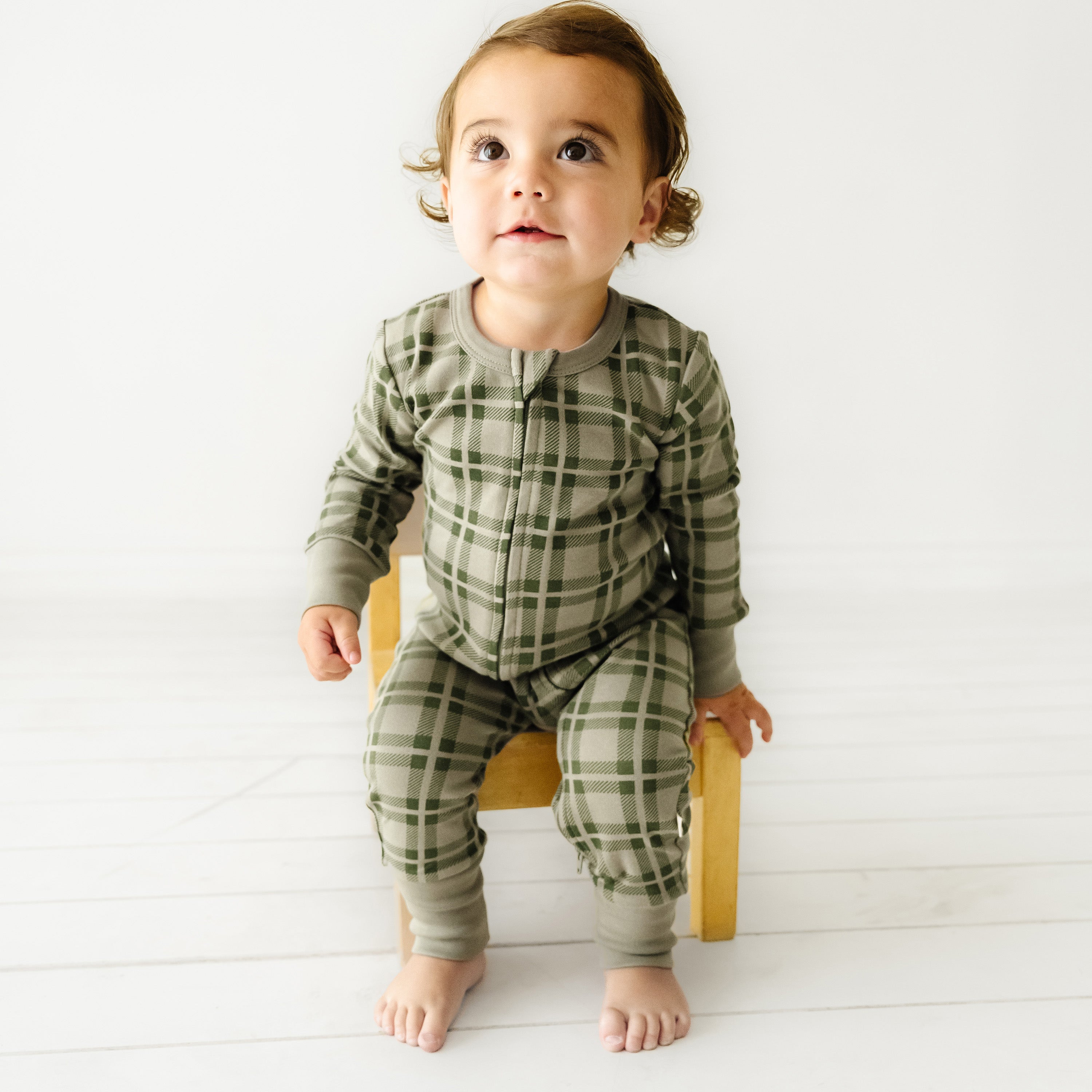 A toddler with curly hair wearing an Organic Baby Organic 2-Way Zip Romper - Plaid sits on a small wooden stool in a bright room.
