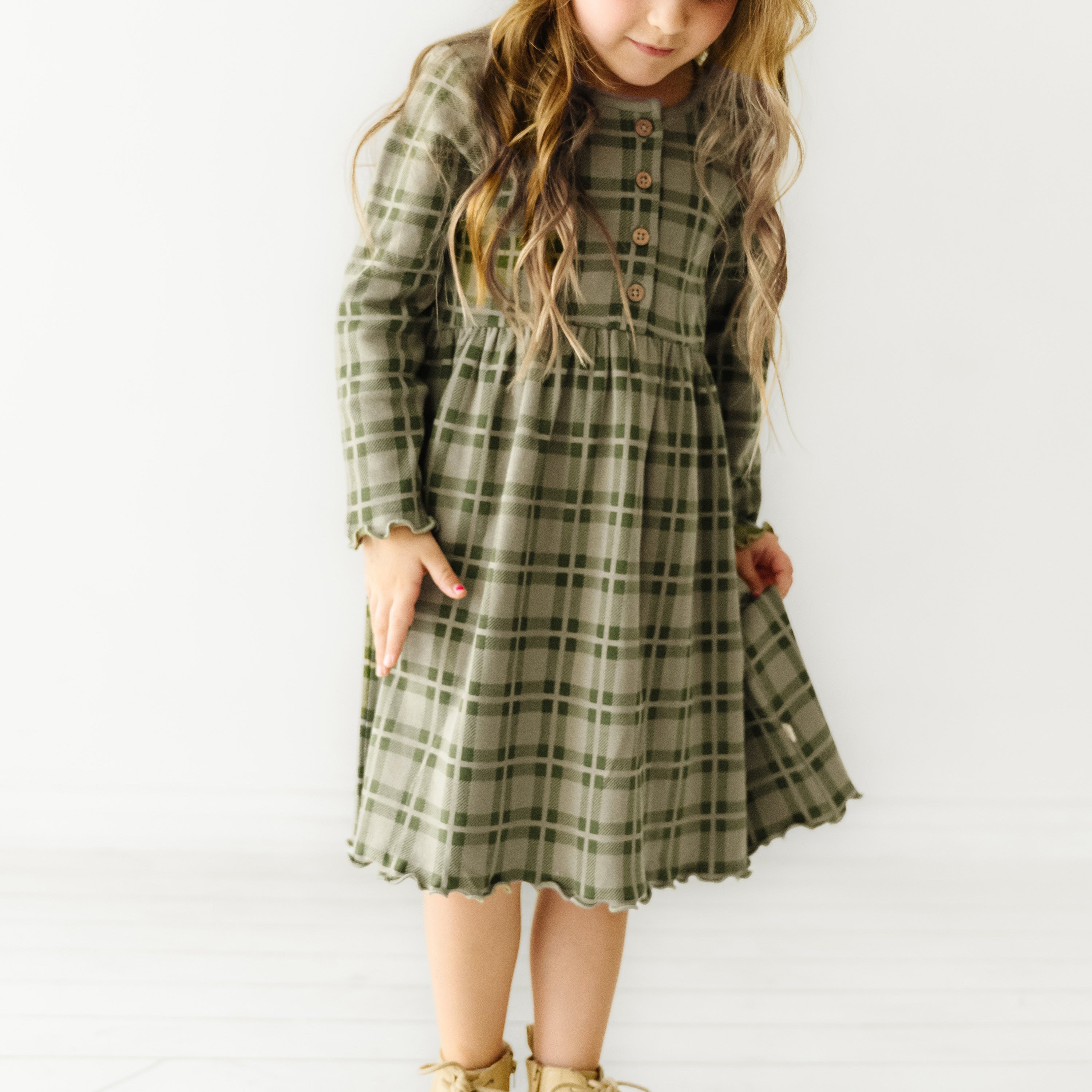 A young girl in an Organic Baby plaid green dress stands confidently, her hair in loose curls, in a bright room with a white background.