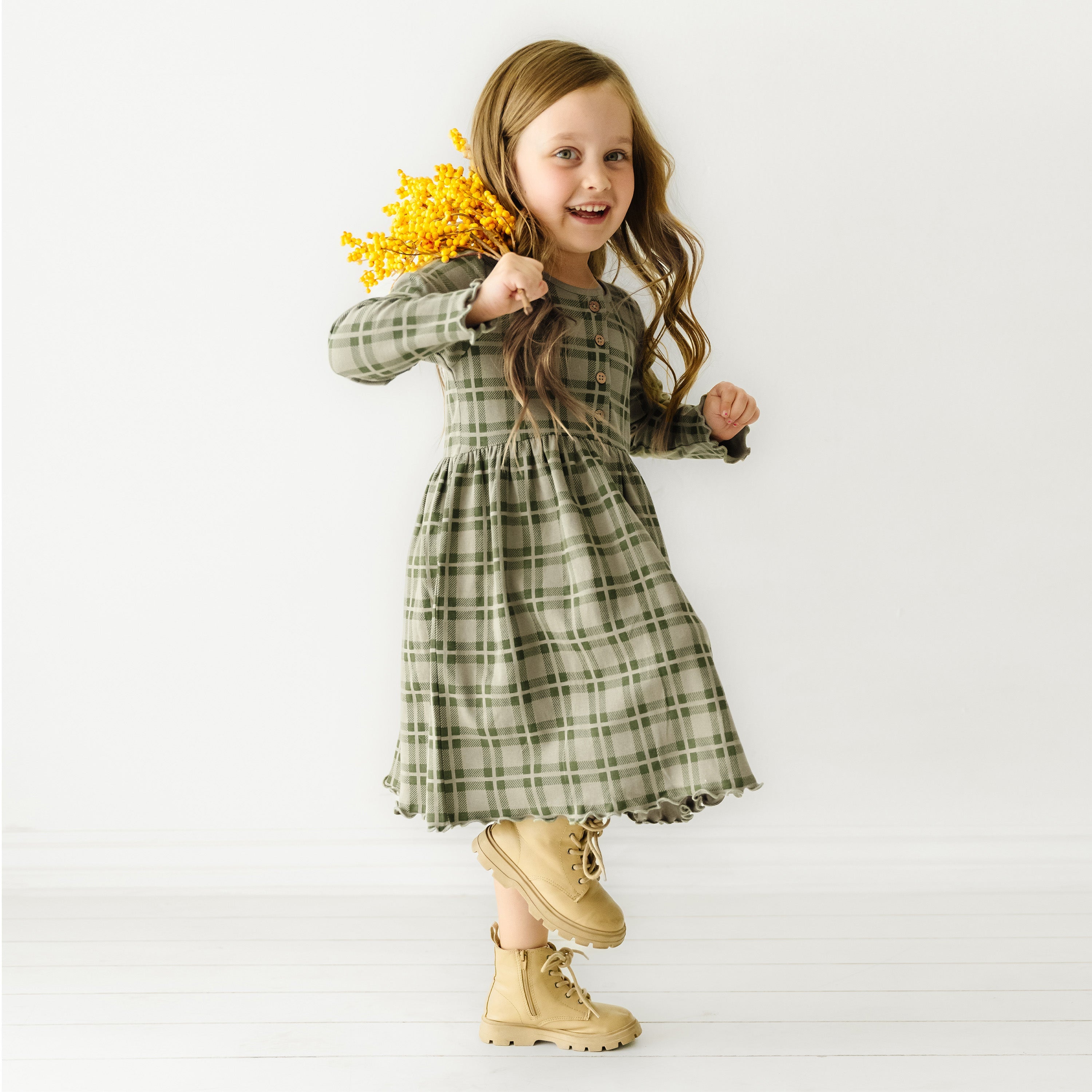A young girl with long hair in a green plaid Organic Baby dress holds yellow flowers, smiling and appearing to run or jump against a plain white background.