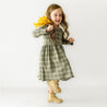 A young girl with long hair in a green plaid Organic Baby dress holds yellow flowers, smiling and appearing to run or jump against a plain white background.