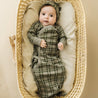 A baby wearing an Organic Baby green plaid kimono knotted sleep gown and matching hat lies in a woven basket, looking upwards with big eyes. The background is soft and neutral.
