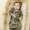 A baby in a green and brown plaid Organic Kimono Knotted Sleep Gown from Organic Baby lies in a woven basket, gazing upwards with wide eyes, a matching hat on its head. The basket is lined
