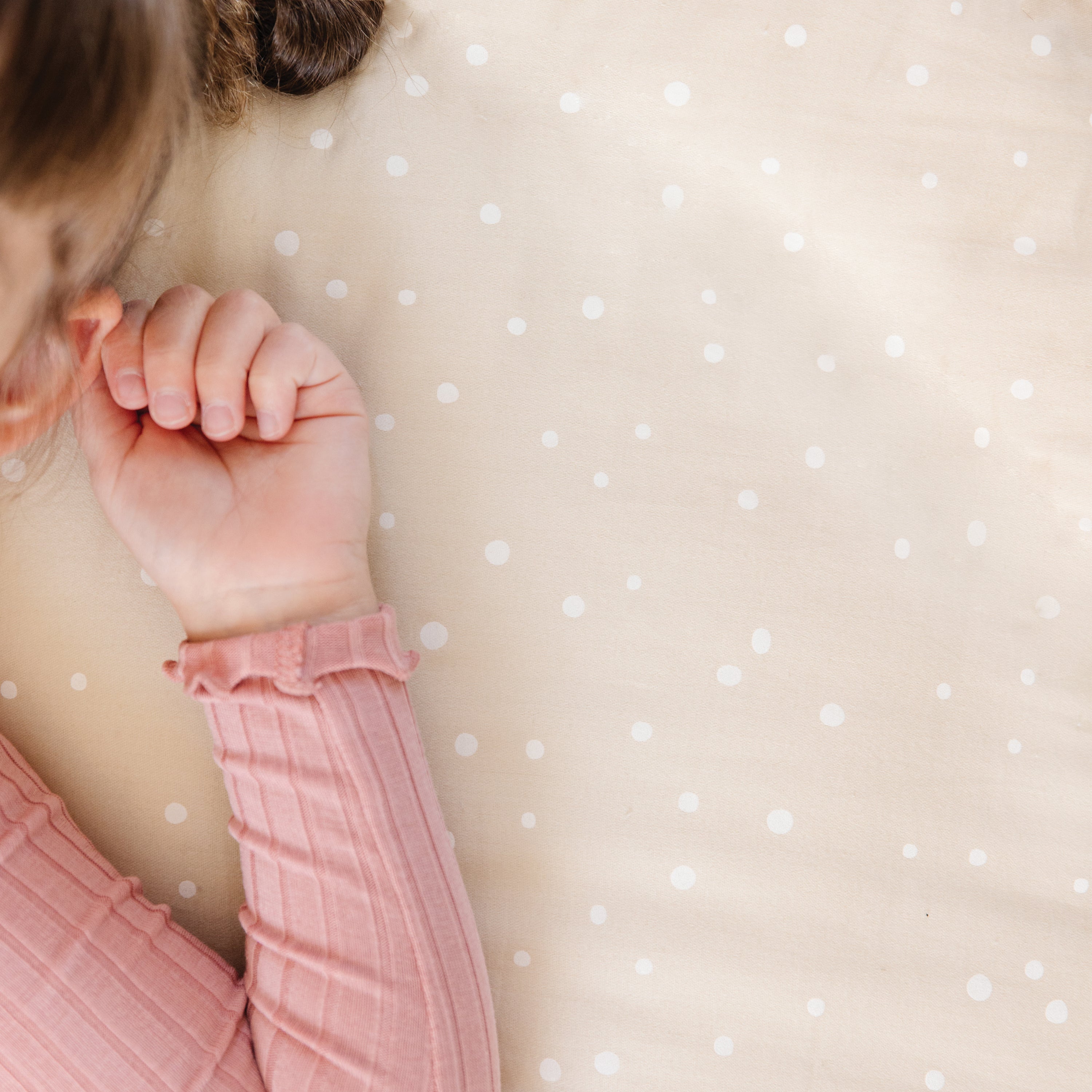 A young child in a pink outfit is lying face down on a Makemake Organics Mini Crib Fitted Sheet in white polka dots, covering her eyes with her hand, showing only her hair and arm.