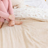 A person sits on a bed grasping their knees, wearing pink pajamas. the bed has floral and Makemake Organics Organic Cotton Sheet Set with polka dot patterns on the linens. only the person's lower body and legs are visible.