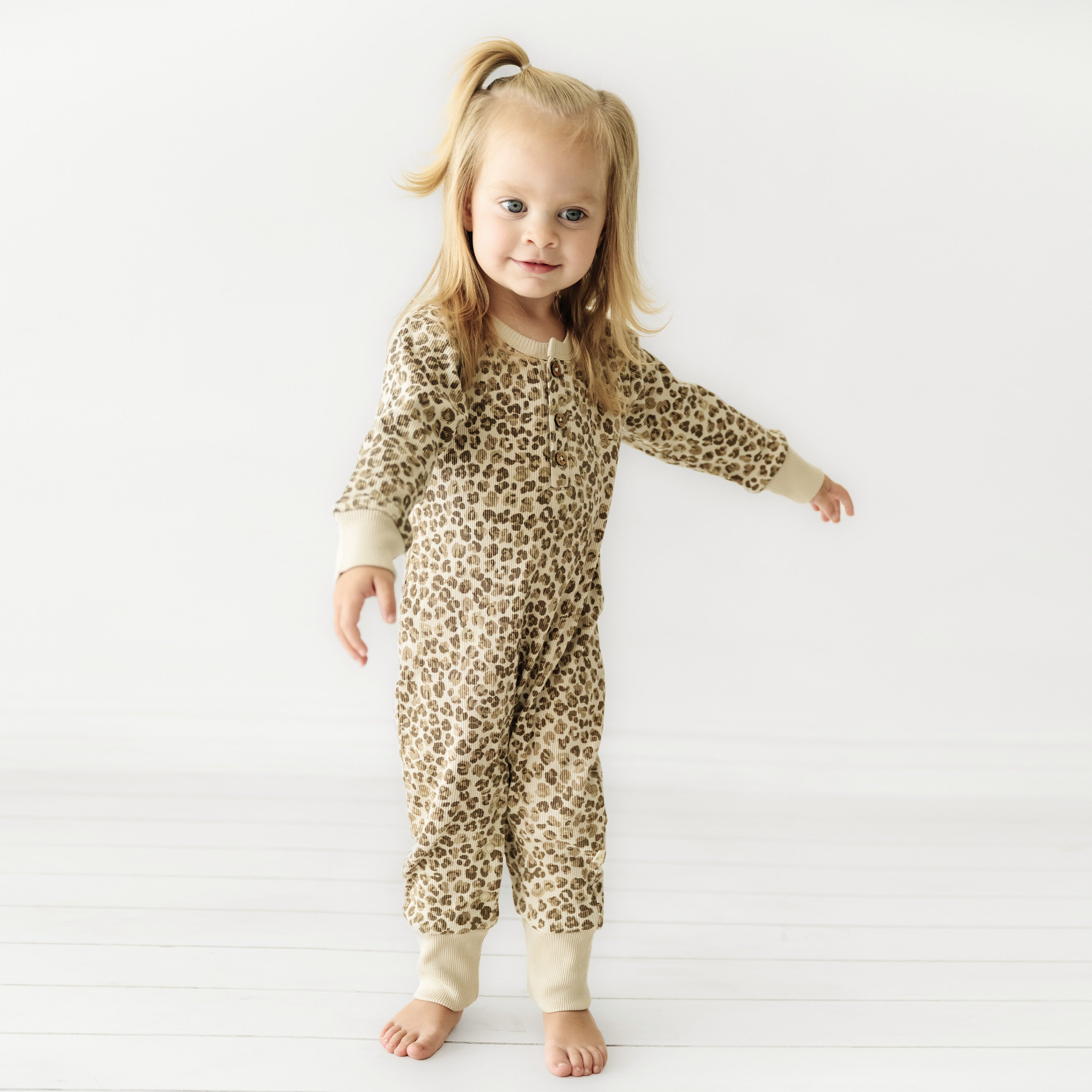 A toddler with blonde hair secured in a ponytail wearing an Organic Baby leopard print jumpsuit stands smiling against a white background.