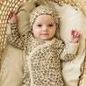 A baby wearing a matching beige outfit from Organic Baby with a heart pattern and a knotted hat lies in a woven basket, looking up with raised arms.