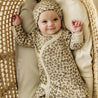 A content baby wearing an Organic Baby Kimono Knotted Sleep Gown in the Spotted pattern and a matching hat lies in a woven bassinet, looking up with a slight smile and raised fists.