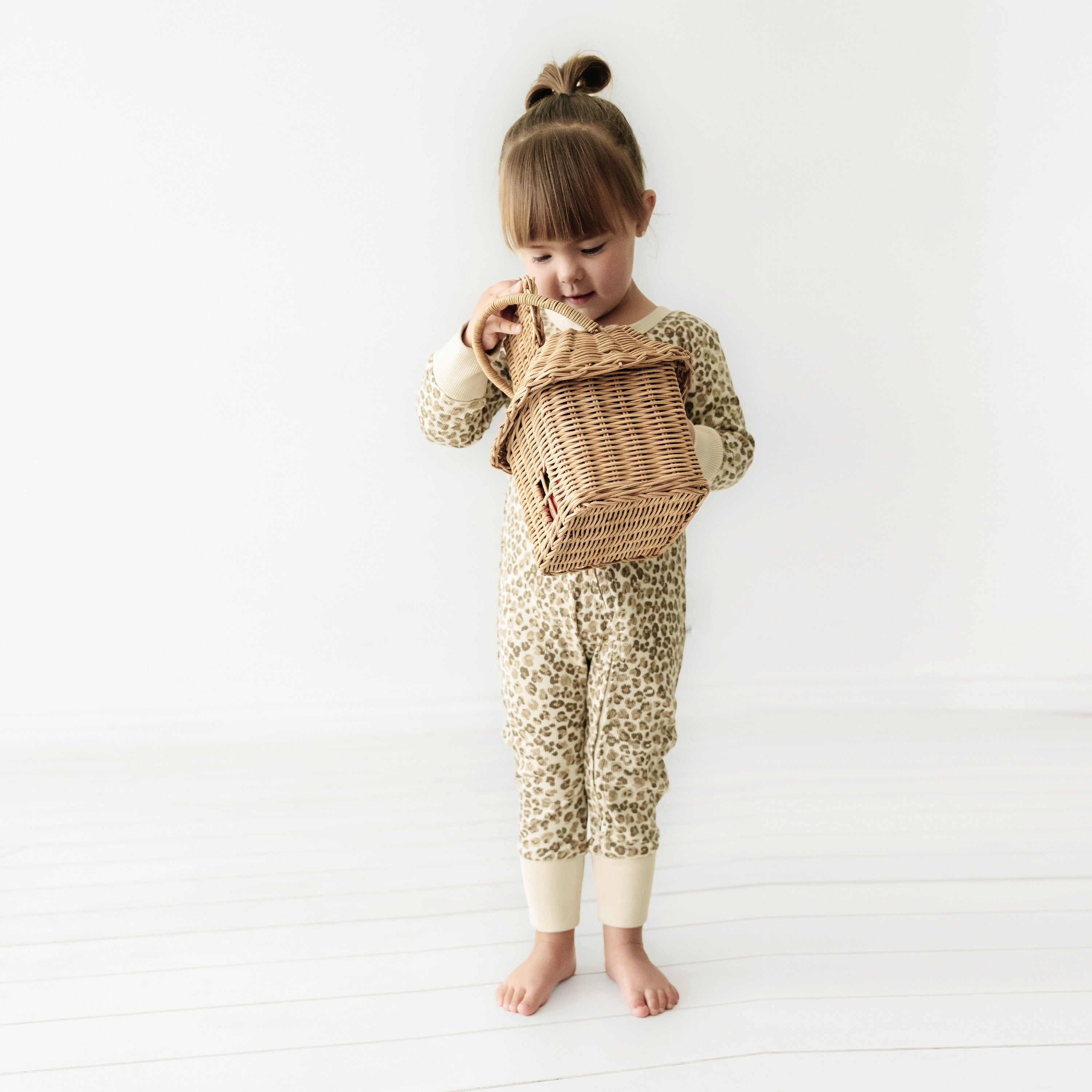 A young child with a cute topknot hairstyle, wearing an Organic Baby leopard print onesie, curiously examines a wicker basket in a bright, white room.