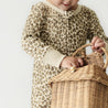 A young child in a beige leopard print dress is handling a wicker basket, seen from a close-up side view. the scene has a soft, minimalist background.