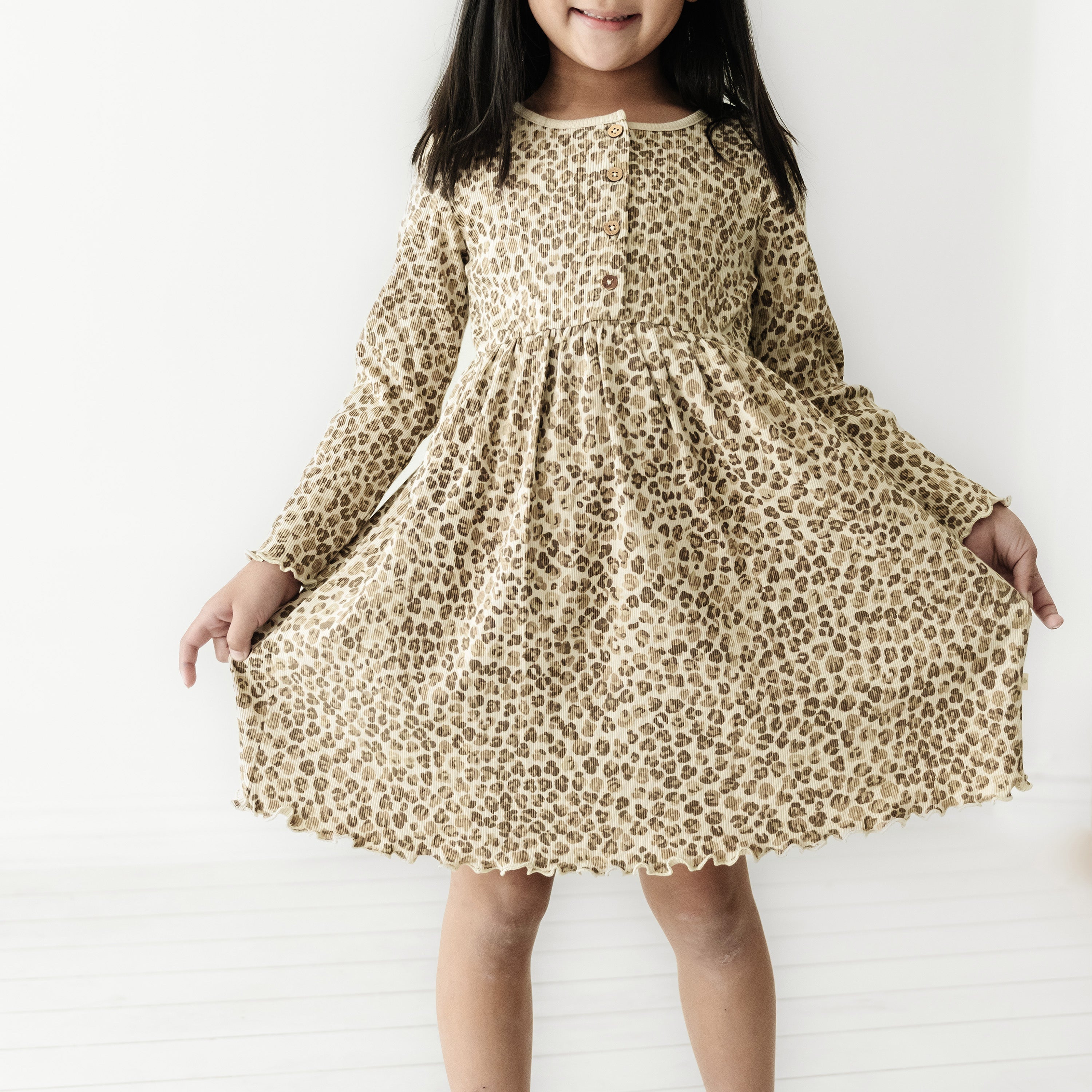 A young girl wearing a flowing Organic Baby leopard print dress, standing against a white background, smiling with her arms slightly outstretched.