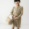 A young girl stands smiling in an Organic Baby leopard print dress, holding a wicker basket bag, against a white background.