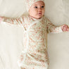 A toddler wearing a Makemake Organics Organic Kimono Knotted Sleep Gown - Summer Floral and matching hat lies on a cream-colored bedspread, gazing upward with a curious expression.