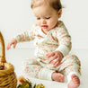 A toddler with light skin and brown hair, dressed in a Makemake Organics Organic Kimono Top & Pants Set - Summer Floral, reaches towards a wicker basket, sitting on a white surface.