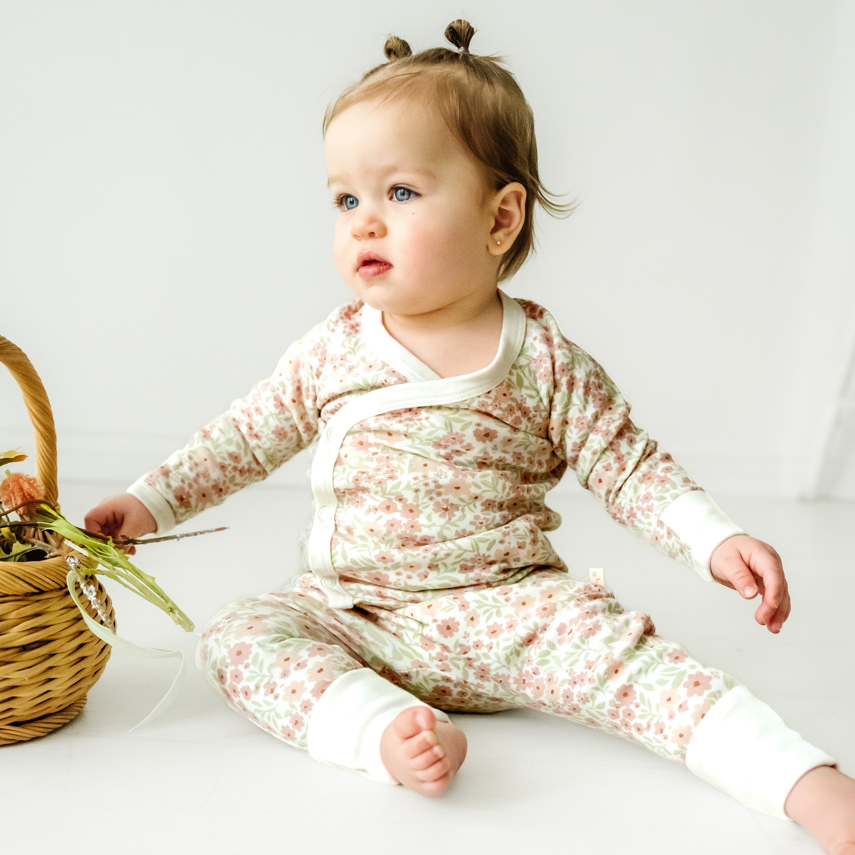 A toddler with two small tufts of hair sits on the floor, wearing a Makemake Organics Organic Kimono Top & Pants Set - Summer Floral, next to a wicker basket, looking to the side with a curious expression.